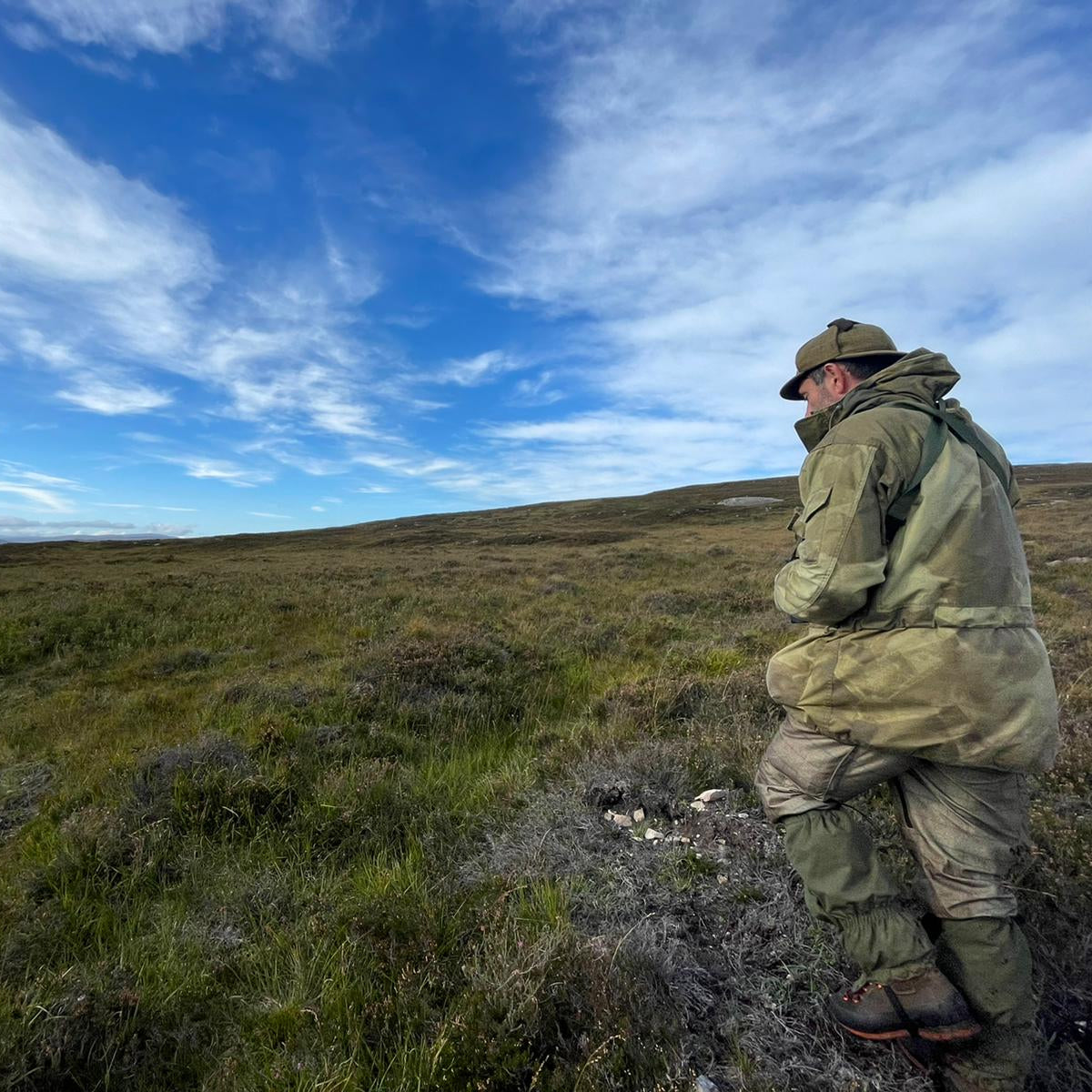 Person in outdoor gear standing on a grassy hillside with a blue sky and clouds.